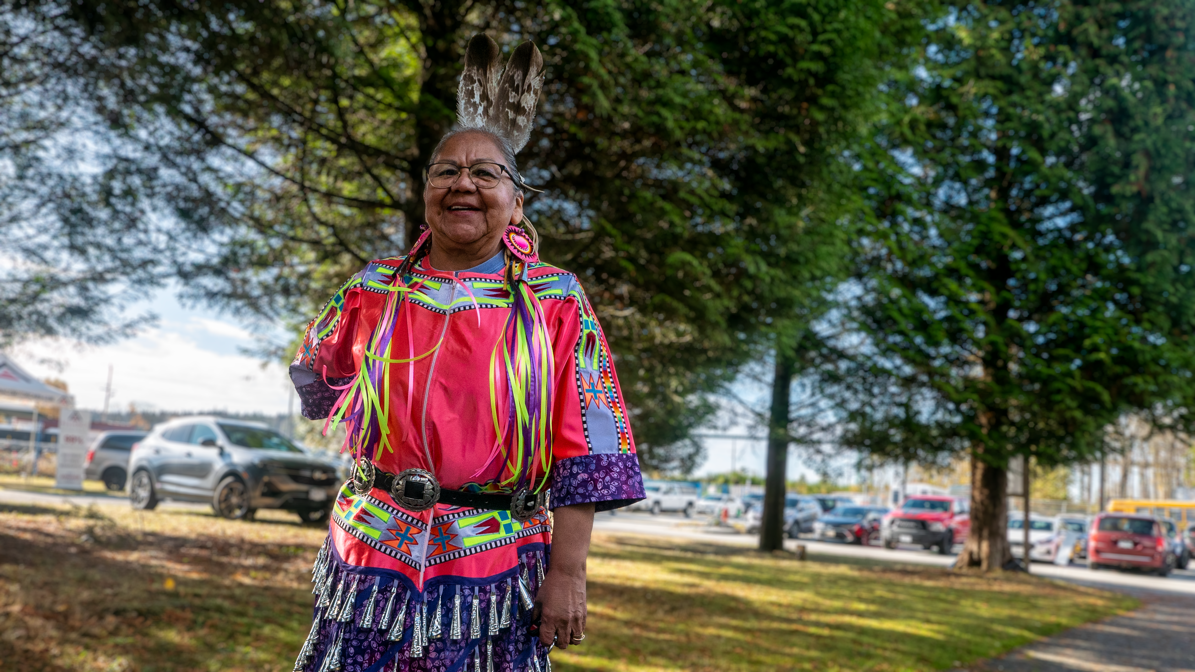 A woman in a multicoloured powwow jingle dress smiles at the camera. She is outdoors in daylight near a parking lot.