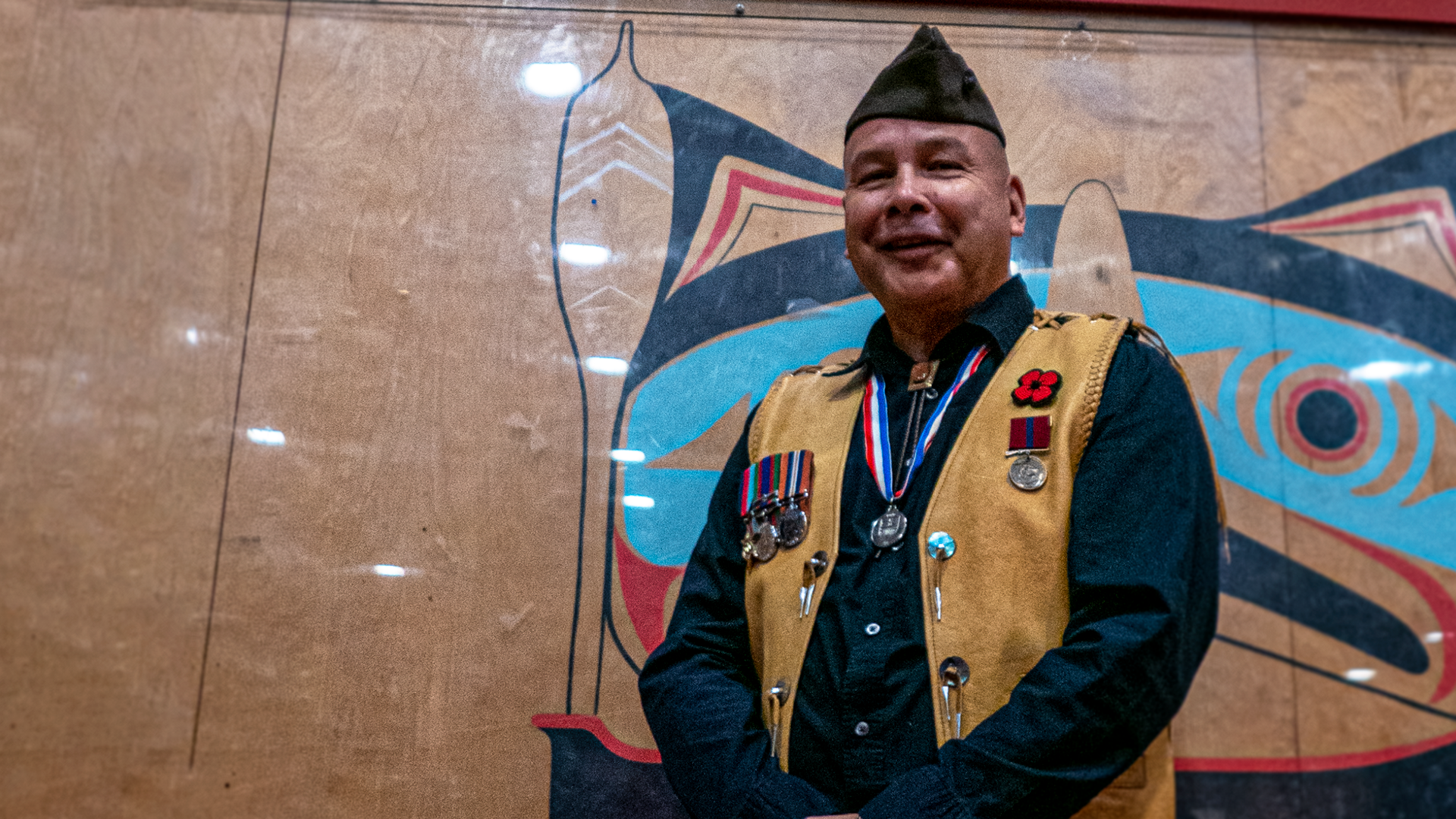 A man in a vest and military cap smiles at the camera, standing in front of a community centre wall decorated in Indigenous art. He is wearing military pins and medals and a poppy pin over his heart.
