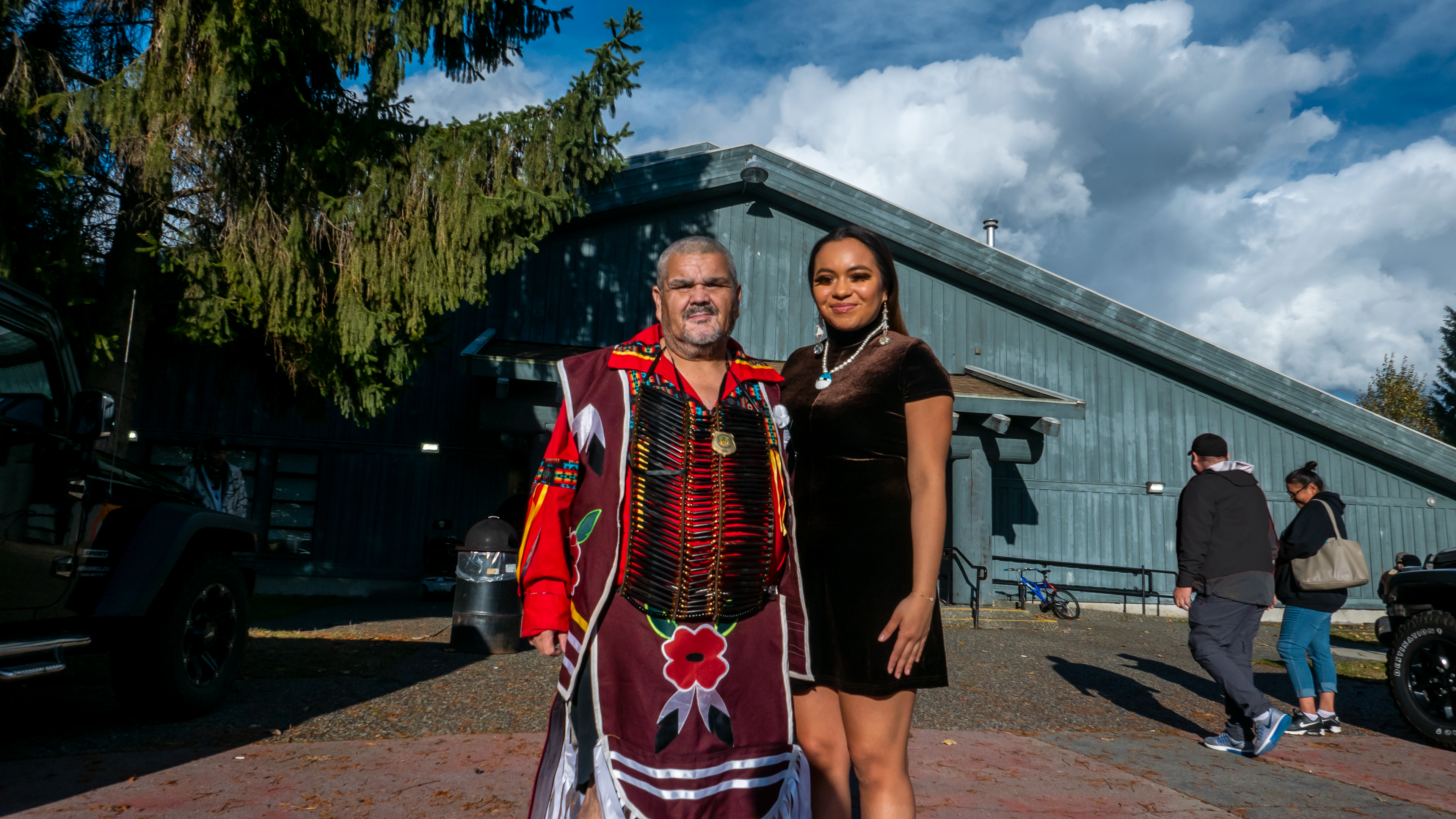 A man wearing traditional Indigenous powwow dressings adorned with poppies stands beside his adult daughter wearing a black dress. They are outdoors in daylight in front of a community centre.