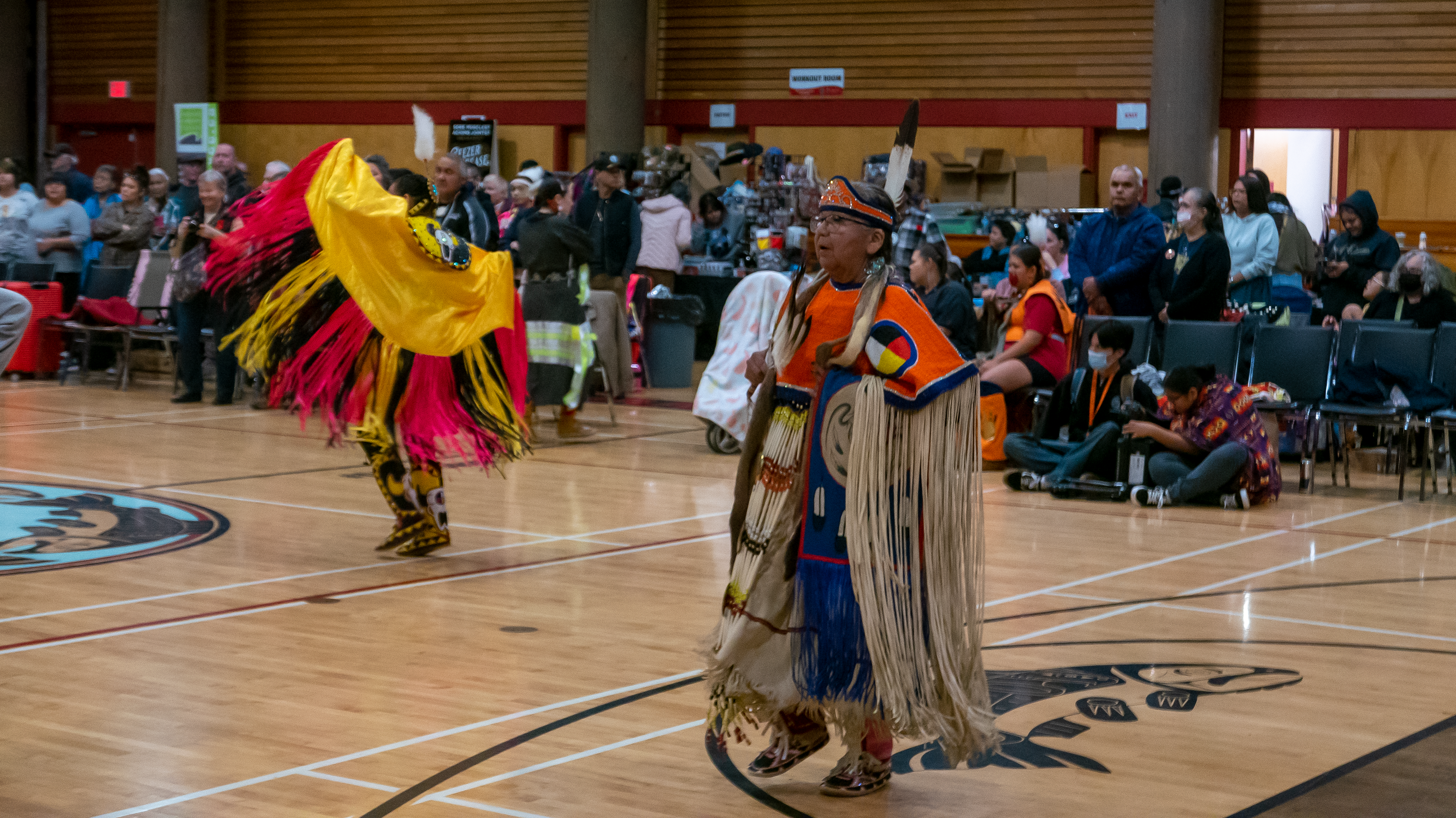 A woman in an orange powwow dress dances inside a crowded community centre. Another powwow dancer spins behind her.
